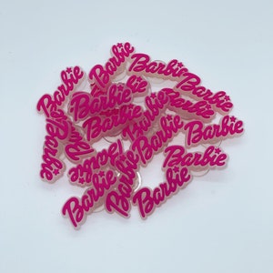 May include: A collection of pink acrylic charms with the word "Barbie" in a cursive font. The charms are scattered on a white surface. The word "Barbie" is in a bright pink color.