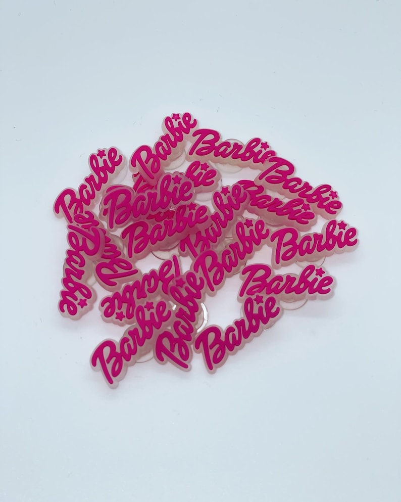 May include: A collection of pink acrylic charms with the word "Barbie" in a cursive font. The charms are scattered on a white surface. The word "Barbie" is in a bright pink color.