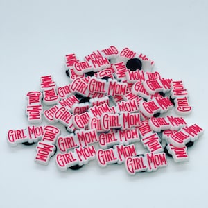 May include: A pile of white shoe charms with the words "GIRL MOM" in red block letters. The charms have a black backing and are scattered on a white surface. The image is well-lit and the charms are in focus.