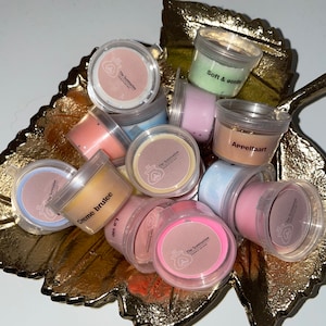 Op de afbeelding: Een verzameling kleine, doorzichtige potjes gevuld met kleurrijke wax melts. De potjes zijn gelabeld met verschillende geuren, waaronder "Crème brûlée", "Zacht en zacht", "Appeltaart" en "Het leven".
