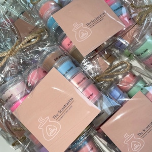 Op de afbeelding: Een selectie van wax melts in kleine, doorzichtige containers, verpakt in doorzichtige plastic zakken. De wax melts zijn verkrijgbaar in verschillende kleuren en geuren, waaronder "Fresh Linen", "Missy Dior", "La Vie" en "GG Guilty". De zakken zijn vastgebonden met touw en voorzien van het merknaam "The Scentsation" en de slogan "Waxmelts and More".