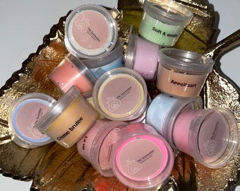 Op de afbeelding: Een verzameling kleine, ronde, doorzichtige containers gevuld met kleurrijke wax melts. De containers hebben labels met de tekst "The Scentstation" en verschillende geuren, waaronder "Soft & Gentle", "Appeltaart", "Crème brûlée" en "La Vie en Rose".