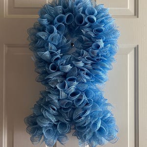 May include: A light blue mesh ribbon wreath in a figure-eight shape, hanging on a white door. The wreath is made of tightly woven, rolled mesh, creating a full, textured appearance. The color is a soft, pastel blue, with a slight shimmer.