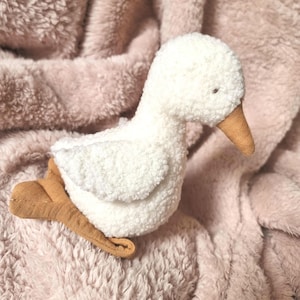 May include: A white plush goose toy with a brown felt beak and feet. The goose is lying on a pink fuzzy blanket.