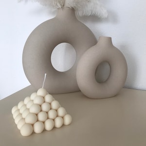 Puede incluir: Dos jarrones de cerámica beige con una abertura en forma de rosquilla, uno más grande que el otro, con un penacho de plumas blancas en el jarrón más grande. Una vela blanca en forma de pirámide con una superficie texturizada se encuentra delante de los jarrones.
