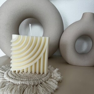 Peut inclure: Une bougie carrée crème avec un motif géométrique en couches, posée sur un dessous de verre en macramé beige. Deux vases beiges en forme de donut sont en arrière-plan. La bougie a une seule mèche blanche.