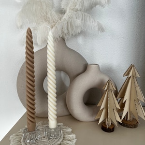 Könnte beinhalten: Zwei gedrehte Kerzen, eine braune und eine weiße, werden auf einem weißen Makramee-Untersetzer präsentiert. Die Kerzen stehen in Glashaltern. Zwei kleine Holz-Weihnachtsbäume mit goldenen Glitzer stehen auf einer hellbraunen Oberfläche. Zwei weiße Federn befinden sich im Hintergrund.