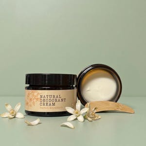 Op de afbeelding: Een donker amberkleurige glazen pot met natuurlijke deodorantcrème, een zwarte deksel en een beige etiket met de tekst Natural Deodorant Cream neroli blossom. Daarnaast staat een open pot met witte crème, een houten spatel en witte nerolibloemen.
