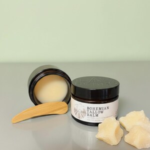 Puede incluir: Dos frascos de color marrón oscuro de bálsamo de sebo Bohemian Tallow Balm, uno abierto y otro cerrado, con una espátula de madera y tres trozos de sebo crudo. La etiqueta presenta un gráfico de montaña y el texto Bohemian Tallow Balm.