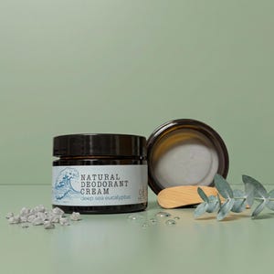 Op de afbeelding: Een donker amberkleurige glazen pot met natuurlijke deodorantcrème, een zwarte deksel en een wit etiket met een blauwe golfillustratie en de tekst Natural Deodorant Cream Deep Sea Eucalyptus. Inclusief een houten spatel en eucalyptus.