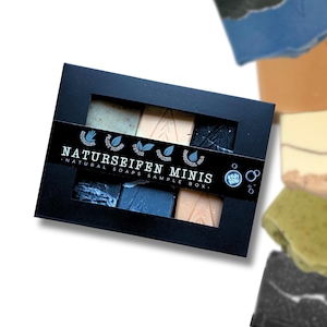 Può includere: Una scatola nera con una finestra trasparente che mostra cinque piccole saponette naturali. La scatola ha un'etichetta che recita "Naturseifen Minis - Natural Soaps Sample Box".