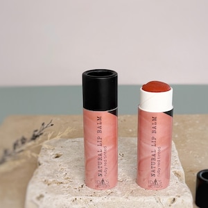 May include: Two tubes of natural lip balm, one with the cap on and the other with the ruby red tinted balm exposed. The tubes are pink with black caps and the text "NATURAL LIP BALM" and "ruby red tinted". The brand name "IDA'S HERBS" is also visible.