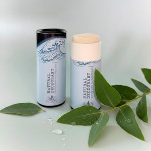 Può includere: Due stick di deodorante naturale con la scritta "Natural Deodorant deep sea eucalyptus" e una grafica a onde. Gli stick sono azzurri e bianchi, con uno stick di deodorante beige visibile. Foglie di eucalipto e goccioline d'acqua in primo piano.