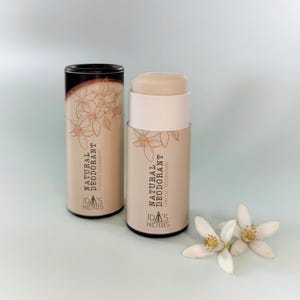 Puede incluir: Dos envases cilíndricos de desodorante natural, uno abierto y otro cerrado, con el texto "NATURAL DEODORANT neroli blossom" y "IDA'S HERBS". El envase abierto muestra el desodorante en barra. Flores blancas de neroli en la parte inferior.