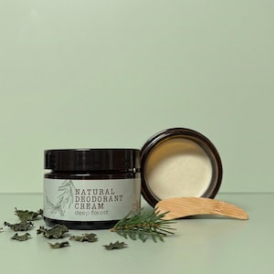 Könnte beinhalten: Ein dunkelbraunes Glasgefäß mit natürlicher Deocreme, beschriftet mit Deep Forest und schwarzem Deckel, dazu ein offenes Gefäß mit weißer Creme, ein Holzspatel, ein kleiner Tannenzweig und verstreute getrocknete grüne Blätter.