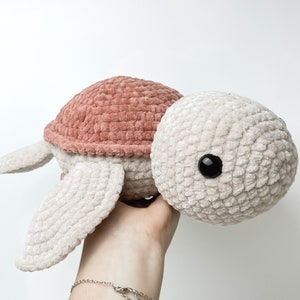 Pu&ograve; includere: Un giocattolo di tartaruga di peluche lavorato a uncinetto con un guscio rosa e un corpo beige. La tartaruga ha occhi neri e una texture morbida e pelosa.