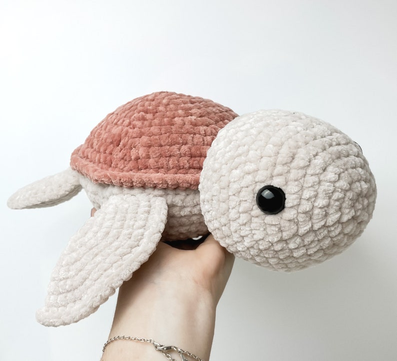 Pu&ograve; includere: Un giocattolo di tartaruga di peluche lavorato a uncinetto con un guscio rosa e un corpo beige. La tartaruga ha occhi neri e una texture morbida e pelosa.