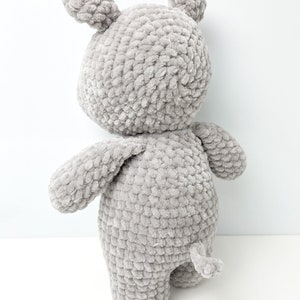 May include: A grey crocheted stuffed animal shaped like a bear. The bear has a long tail and is standing on a white surface.