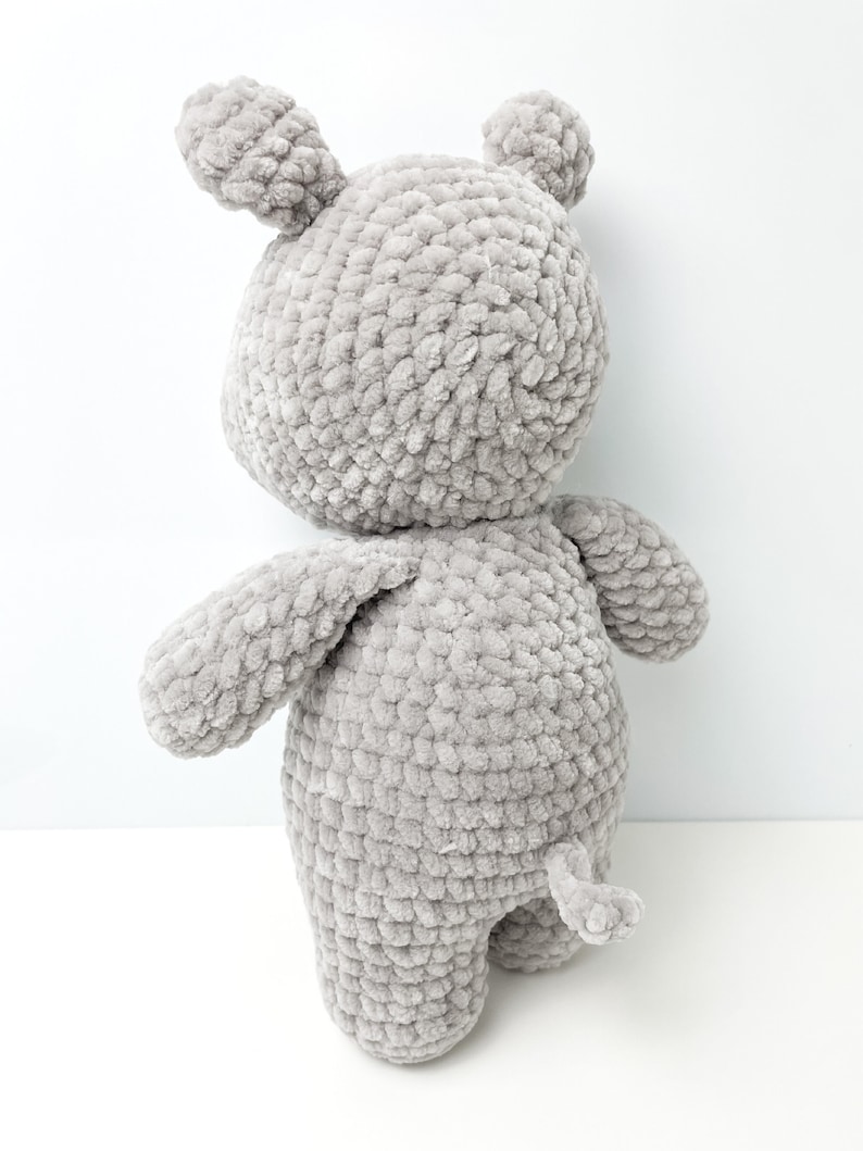 May include: A grey crocheted stuffed animal shaped like a bear. The bear has a long tail and is standing on a white surface.