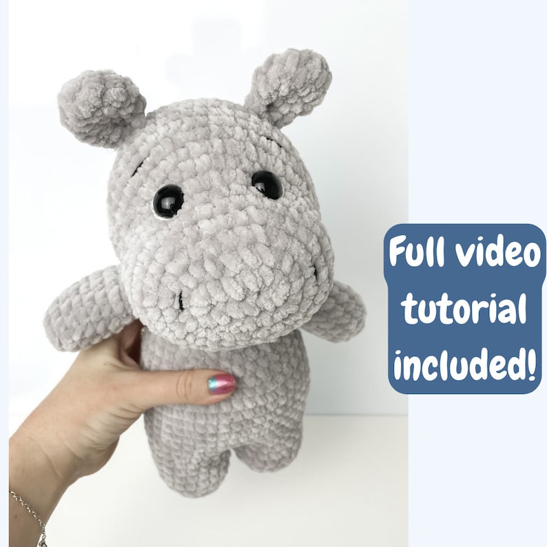 May include: A grey crocheted plush hippopotamus toy with black eyes. The toy is being held by a hand. The text "Full video tutorial included!" is in a blue box in the background.