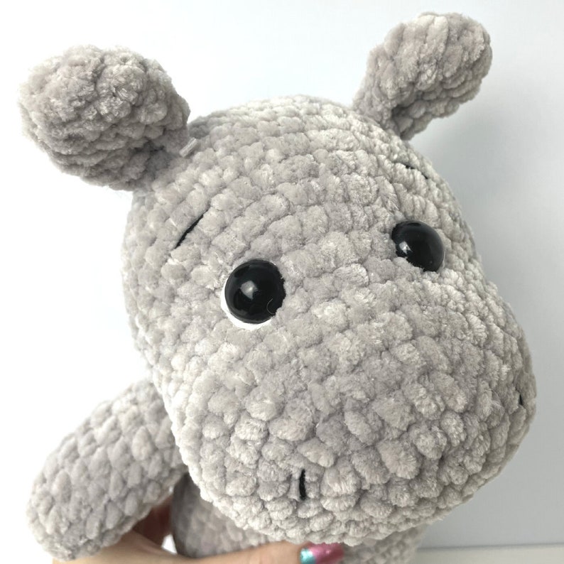 May include: A grey crocheted plush hippopotamus toy with black eyes. The hippopotamus has a textured, fuzzy surface.