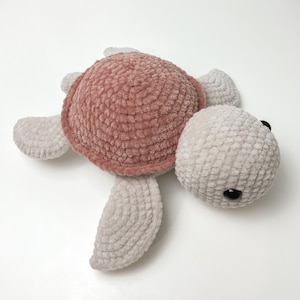 Pu&ograve; includere: Un giocattolo di peluche a forma di tartaruga all'uncinetto con un guscio rosa e arti grigi. La tartaruga ha occhi neri.