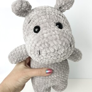 May include: A grey crocheted plush hippopotamus toy with black eyes. The toy is being held by a hand with colourful nail polish.