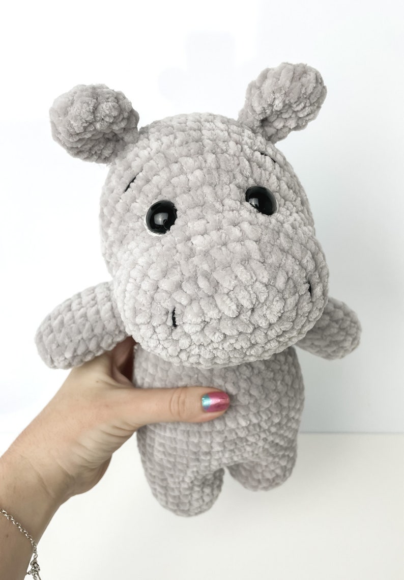 May include: A grey crocheted plush hippopotamus toy with black eyes. The toy is being held by a hand with colourful nail polish.