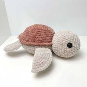 Pu&ograve; includere: Un giocattolo di tartaruga di peluche lavorato a uncinetto con un guscio rosa e un corpo beige. La tartaruga ha un occhio nero e giace su una superficie bianca.