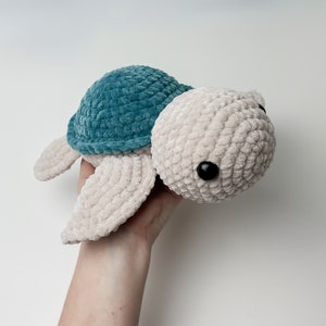May include: A crocheted plush toy turtle with a teal shell and beige body. The turtle has black eyes and a soft, fuzzy texture.