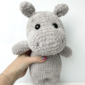 May include: A grey crocheted plush hippopotamus toy with black eyes. The toy is being held by a hand.