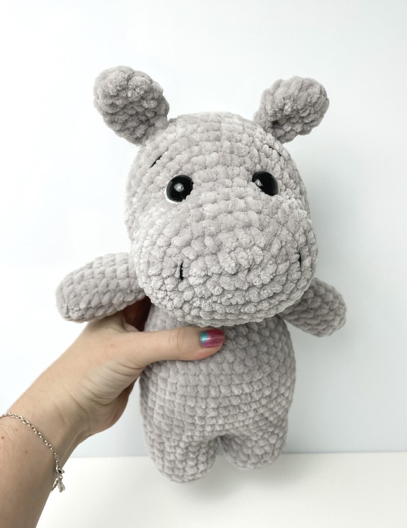May include: A grey crocheted plush hippopotamus toy with black eyes. The toy is being held by a hand.