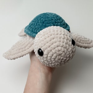 May include: A crocheted plush toy turtle with a beige body and a teal shell. The turtle has black eyes and a friendly expression.