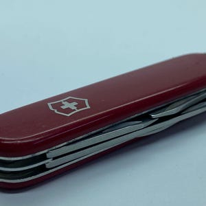 May include: A red Swiss Army knife with multiple stainless steel tools. The knife features a silver emblem with a white cross on a red shield. The tools are partially extended, showcasing their functionality.