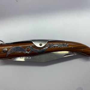 May include: A folding pocket knife with a wooden handle and a stainless steel blade. The handle has a decorative metal inlay with star and crescent designs. The blade is partially open, revealing the word "OKAPI". A metal ring is attached to the end of the handle.