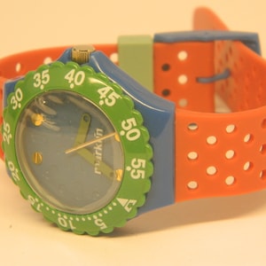 May include: A blue and green wristwatch with a red and orange perforated strap. The watch face has a blue background with white markings and the word "markin" printed on it.