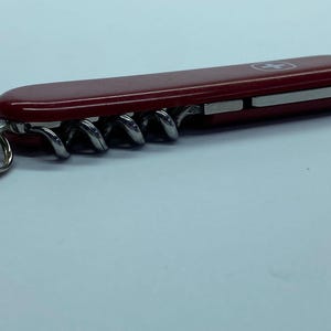 May include: A red Swiss Army knife featuring a silver corkscrew, a small silver key ring, and a silver emblem. The knife is closed, displaying its smooth, glossy red casing. The corkscrew is visible.