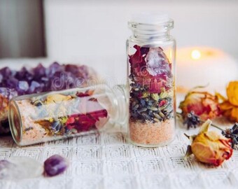 May include: Two small glass bottles filled with layers of pink salt, dried flowers, and herbs. Amethyst crystals and dried roses are arranged around the bottles. A lit candle is in the background, creating a soft, warm ambiance.