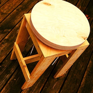 May include: A wooden stool with a round, light wood top. The stool is made of unfinished wood and has a simple, rustic design.