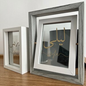 May include: Two white picture frames with gray wood trim. The frames are on a wooden surface. The glass in the frames has gold lettering. The left frame says "Dris" and the right frame says "Layla".