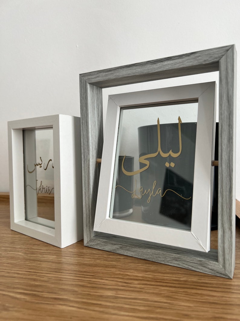 May include: Two white picture frames with gray wood trim. The frames are on a wooden surface. The glass in the frames has gold lettering. The left frame says "Dris" and the right frame says "Layla".