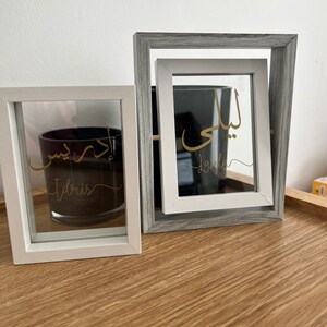 May include: Two white picture frames with glass fronts. The frames are on a wooden surface. The top frame is gray and has the Arabic word "Laila" written in gold inside. The bottom frame has the Arabic word "Idris" written in gold inside.