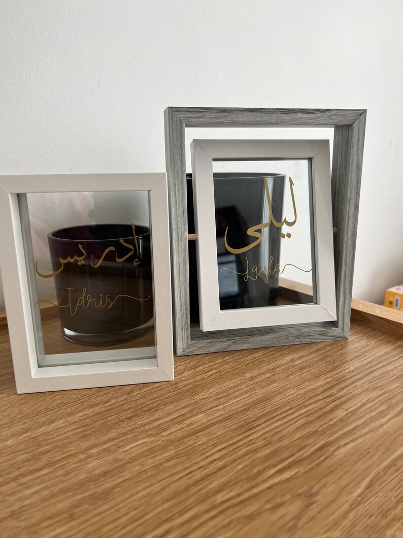 May include: Two white picture frames with glass fronts. The frames are on a wooden surface. The top frame is gray and has the Arabic word "Laila" written in gold inside. The bottom frame has the Arabic word "Idris" written in gold inside.