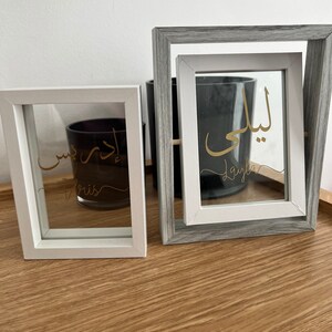 May include: Two white picture frames with glass fronts. The frames are on a wooden surface. The left frame has the Arabic word "Layla" written in gold script. The right frame has the Arabic word "Layla" written in gold script.