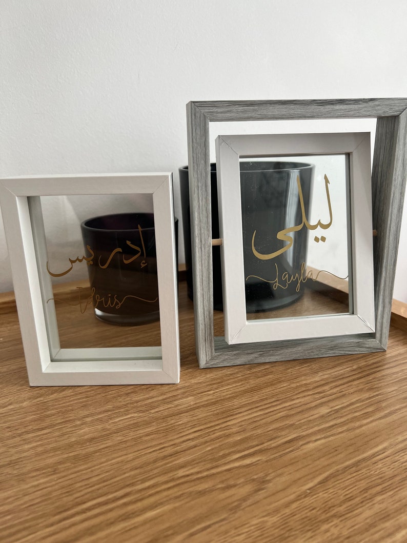 May include: Two white picture frames with glass fronts. The frames are on a wooden surface. The left frame has the Arabic word "Layla" written in gold script. The right frame has the Arabic word "Layla" written in gold script.
