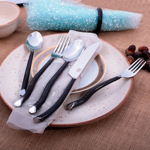 May include: A set of four silver utensils with black, twisted handles. The utensils are arranged on a white plate with a gold rim and a white napkin. The utensils include a fork, a knife, and two spoons.