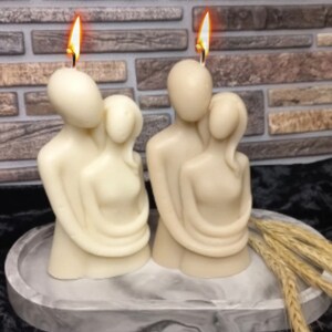Puede incluir: Dos velas esculpidas de color marfil que representan figuras abrazadas, con mechas encendidas. Las velas est&aacute;n sobre una bandeja veteada gris y blanca, con espigas de trigo a un lado. El fondo presenta una pared de ladrillo.