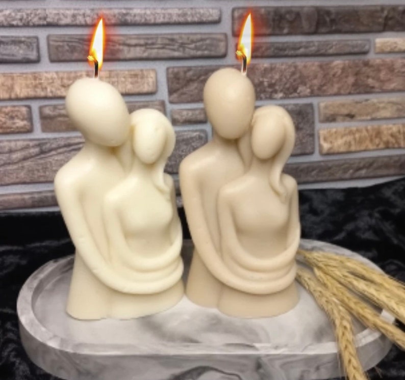 Puede incluir: Dos velas esculpidas de color marfil que representan figuras abrazadas, con mechas encendidas. Las velas est&aacute;n sobre una bandeja veteada gris y blanca, con espigas de trigo a un lado. El fondo presenta una pared de ladrillo.
