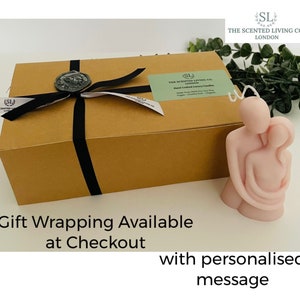 Puede incluir: Una vela rosa claro con la forma de dos figuras abraz&aacute;ndose, junto a una caja de regalo marr&oacute;n atada con una cinta negra y un sello de cera. La caja tiene una etiqueta que dice "The Scented Living Co. London". El texto en la imagen dice "Gift Wrapping Available at Checkout".
