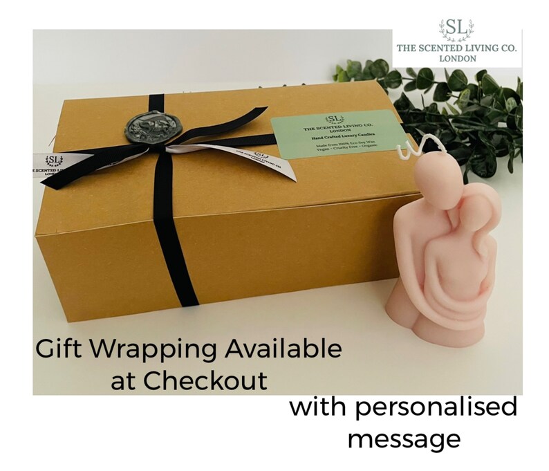 Puede incluir: Una vela rosa claro con la forma de dos figuras abraz&aacute;ndose, junto a una caja de regalo marr&oacute;n atada con una cinta negra y un sello de cera. La caja tiene una etiqueta que dice "The Scented Living Co. London". El texto en la imagen dice "Gift Wrapping Available at Checkout".